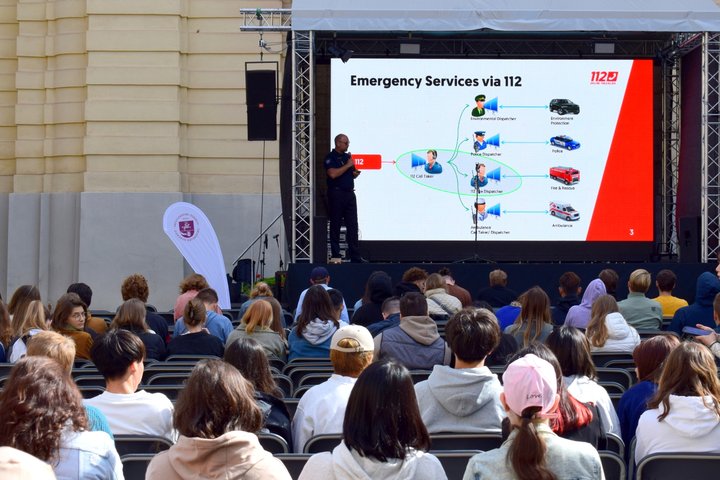Nuotraukoje matoma kieme esanti scena, ekranas ir skaidres komentuojantis pareigūnas.