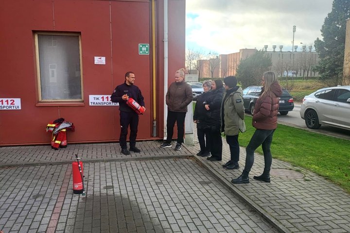 Nuotraukoje matoma grupė žmonių, iš kurių vienas laiko rankose gesintuvą, rodydamas, kaip juo naudotis