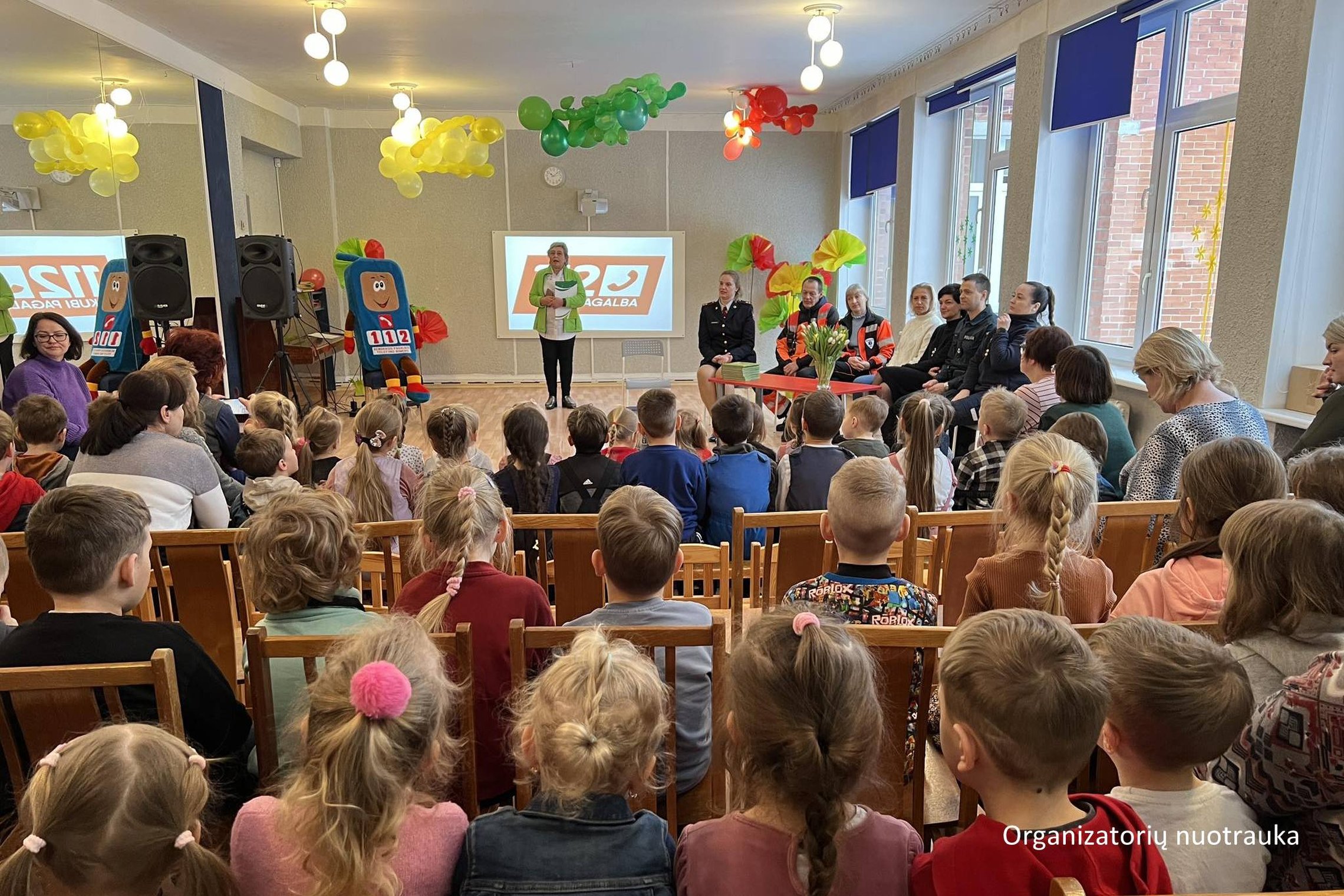 Nuotraukoje matoma vaikų darželio salė, ant kėdučių sėdintys vaikai ir kiti renginio dalyviai prie ilgo stalo