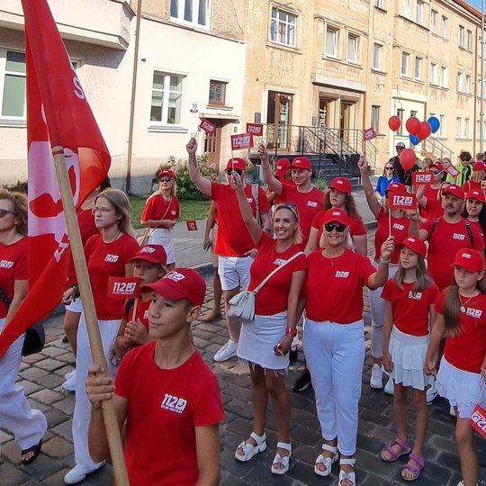Nuotraukoje matomi raudonais marškinėliais vilkintys šventinės eisenos dalyviai, išsirikiavę gatvėje