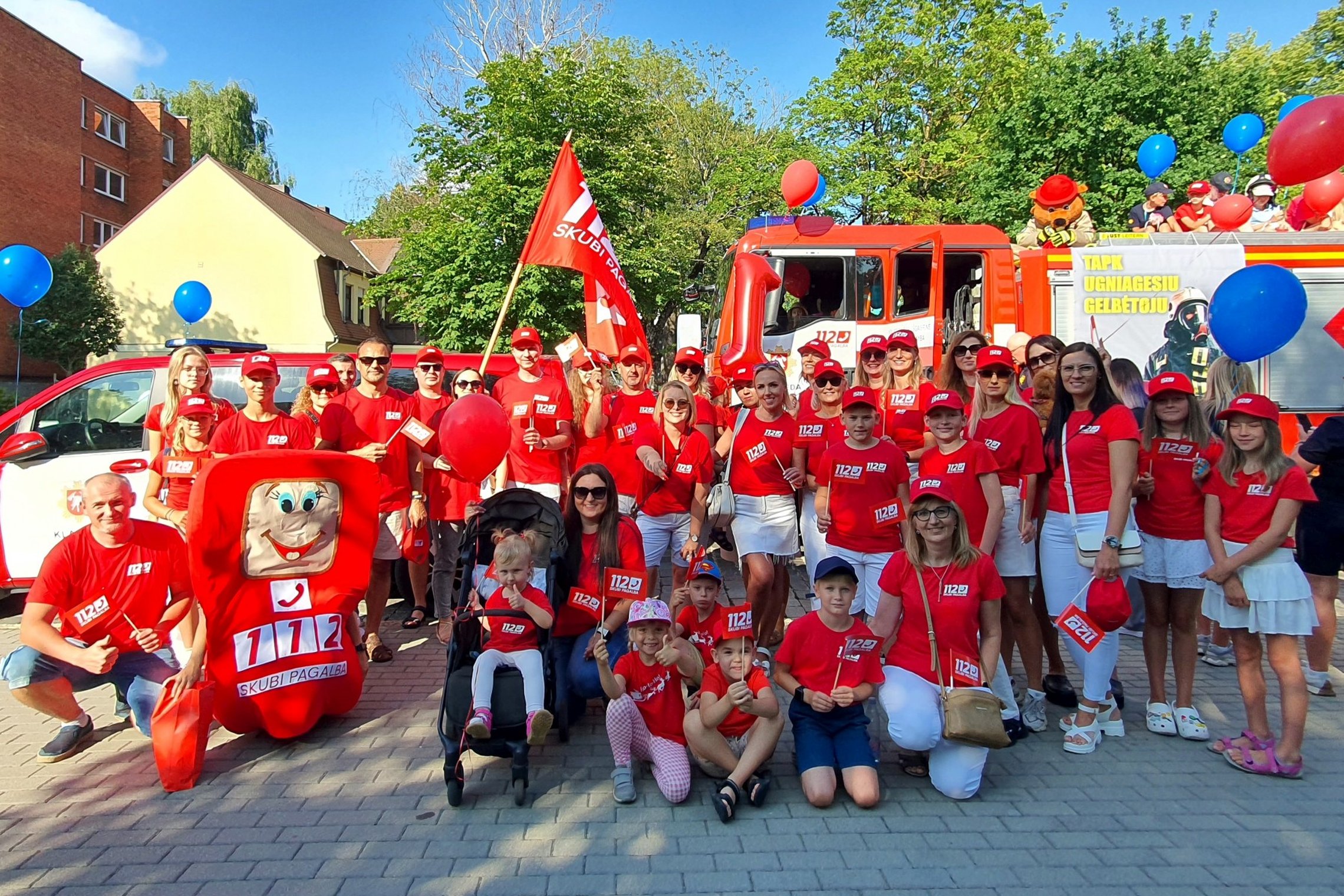 Nuotraukoje matoma didelė grupė raudonais marškinėliais vilkinčių žmonių: vyrų, moterų ir vaikų.