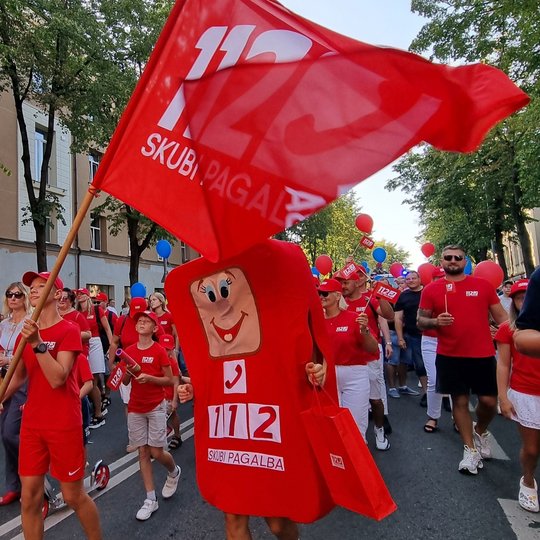 Nuotraukoje matomi  raudonais marškinėliais vilkintys eisenos dalyviai, kurių priekyje eina vėliavą nešantis berniukas ir personažas, vardu Telis