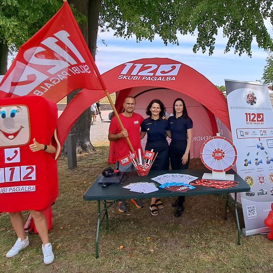 Nuotraukoje matoma palapinė, joje stovintis vyras su vėliava ir dvi moterys. Šalia palapinės stovi ir personažas, vardu Telis