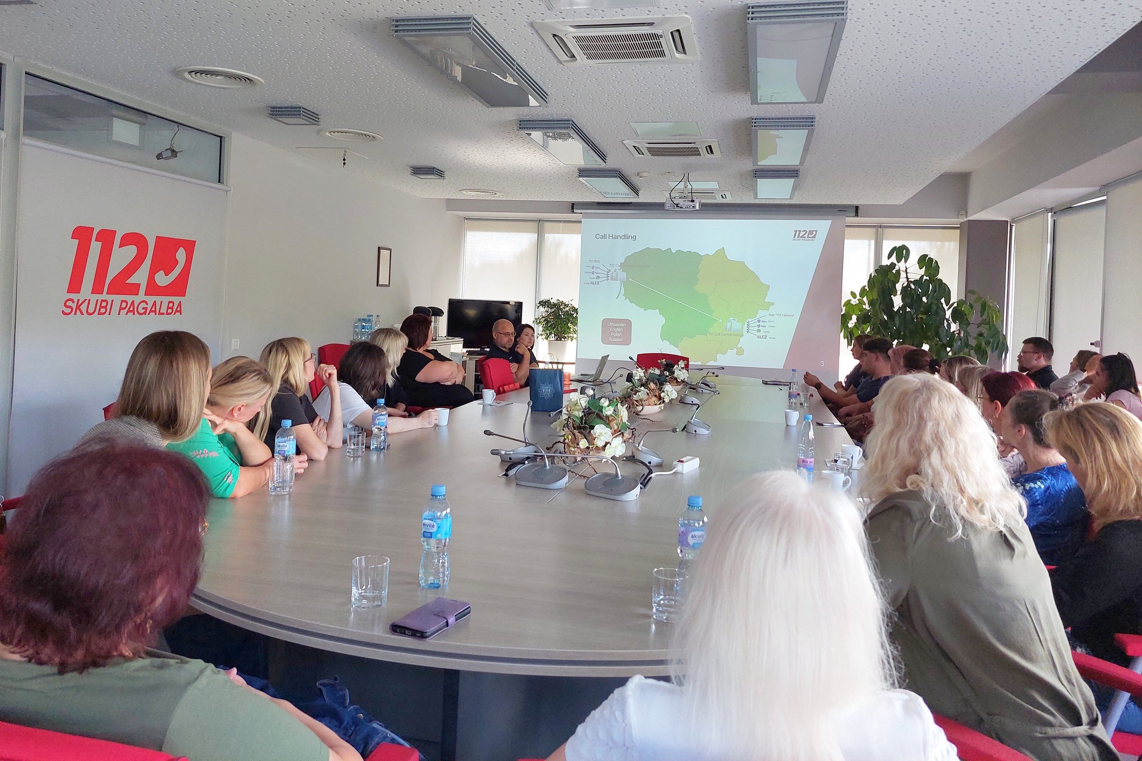 Nuotraukoje matoma posėdžių salė, kurioje aplink stalą sėdi daug žmonių ir ekranas, kuriame demonstruojamos skaidrės apie Bendrąjį pagalbos centrą