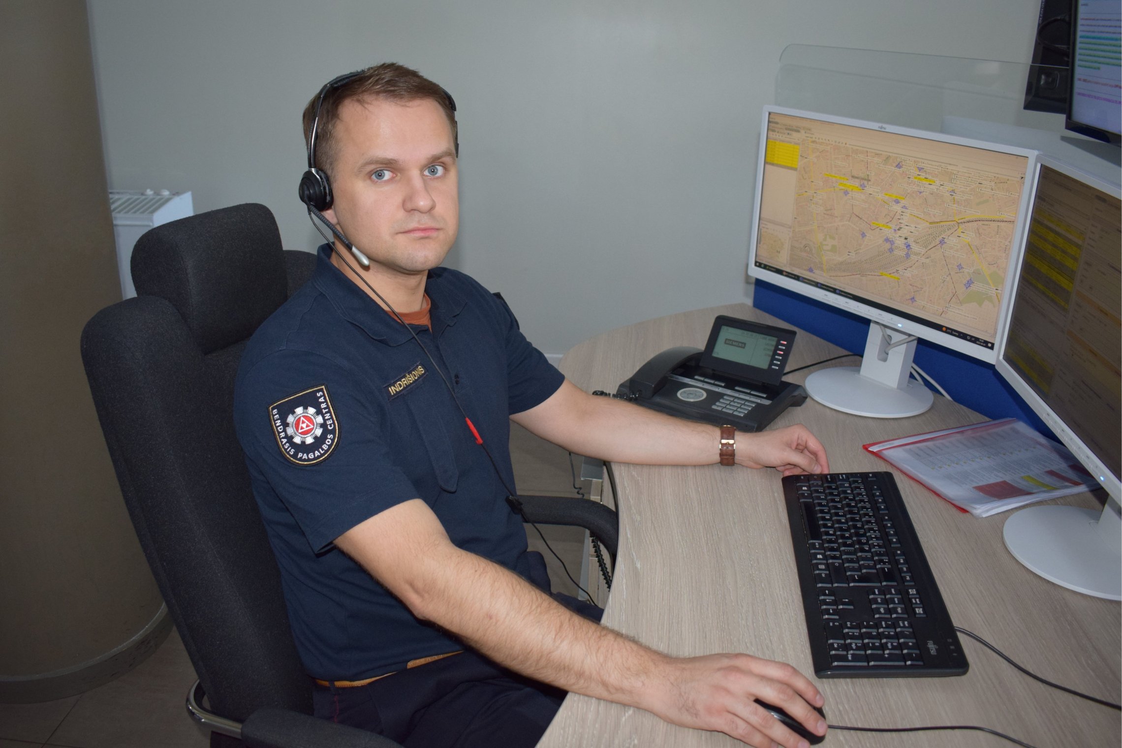 Nuotraukoje matomas operatoriaus apranga vilkintis ir darbo vietoje prieš monitorius sėdintis jaunas vyras