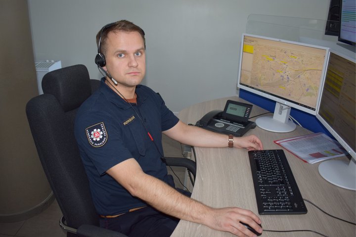 Nuotraukoje matomas operatoriaus apranga vilkintis ir darbo vietoje prieš monitorius sėdintis jaunas vyras