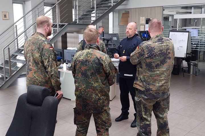 Nuotraukoje matomas Operatorių salėje stovintis Bendrojo pagalbos centro viršininko pavaduotojas ir jį apsupęs pulkelis Vokietijos karių uniformomis vilkinčių žmonių.