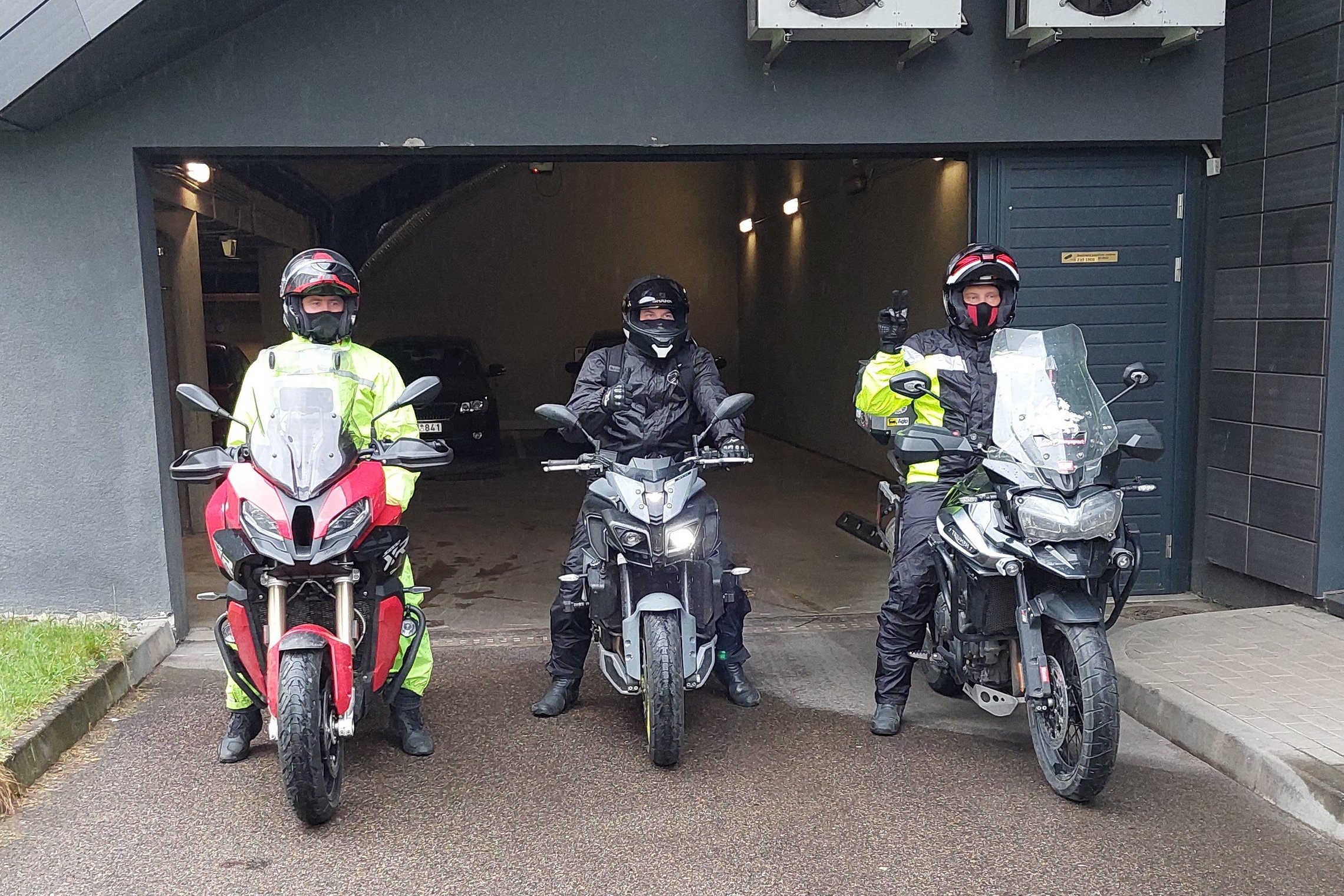 Nuotraukoje matomi trys motociklininkai, sėdintys ant motociklų, prieš pakeltus požeminio garažo vartus 