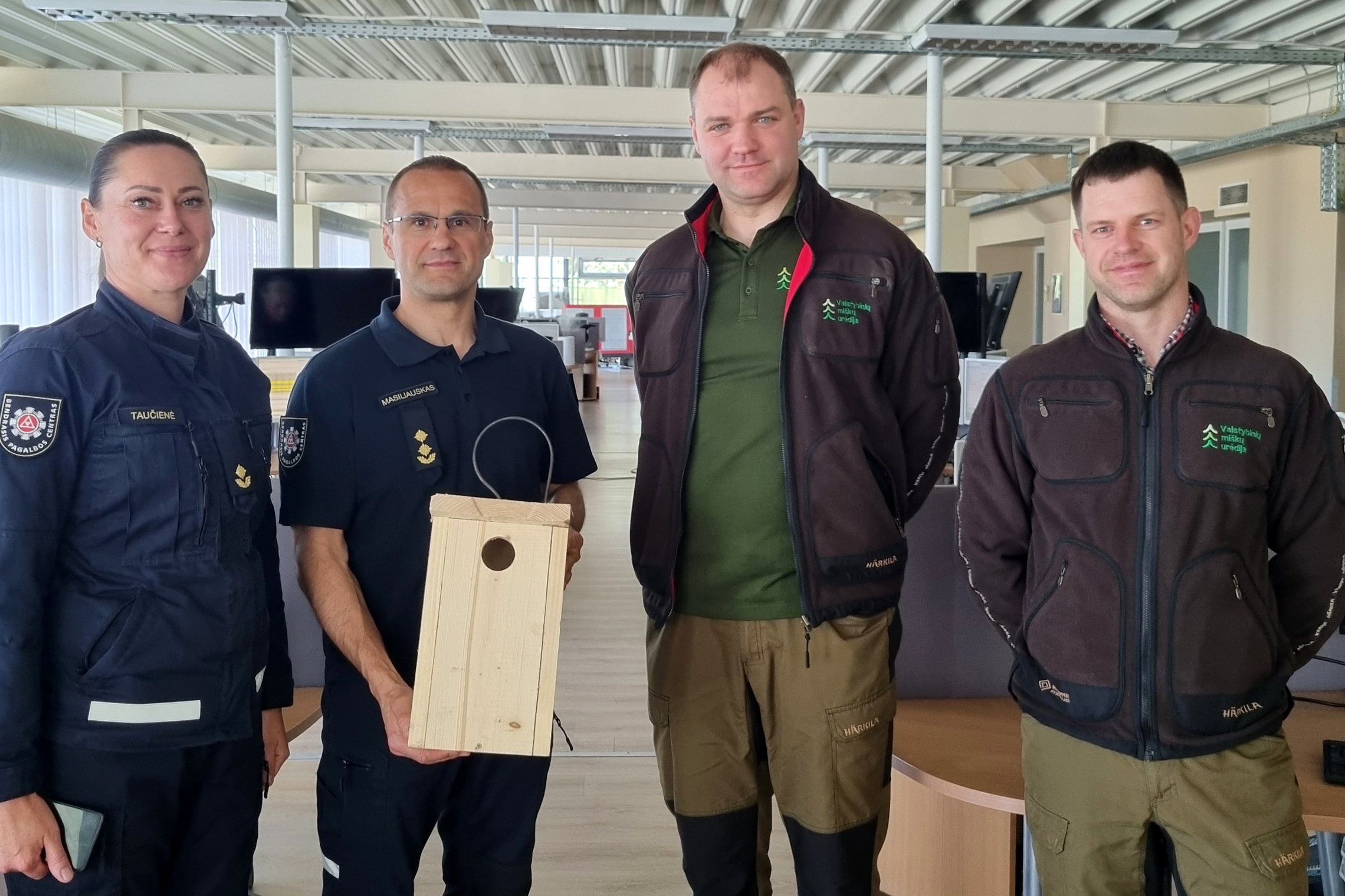 Nuotraukoje matomi keturi žmonės - moteris ir vyras, dirbantys Bendrajame pagalbos centre, ir du miškininkai. Vyras iš Bendrojo pagalbos centro savo rankose laiko inkilą.