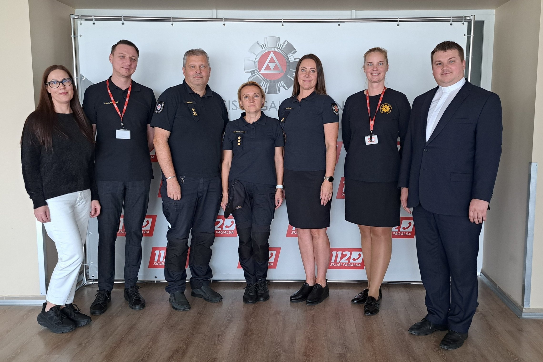 Nuotraukoje matoma prie sienelės išsirikiavusi grupė besišypsančių žmonių